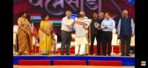 Rambhai Patel is seen getting award from the chief guest with 3 women on the left and 4 men on the tight