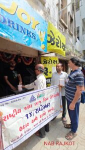 This image shows a group of people standing outside a small shop named "Joy Drinks," as seen from the signboard above the stall. The shop appears to be serving refreshments, and a man behind the counter is seen handing over a donation can to an elderly man in front.