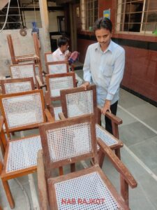 A man, who appears to be visually impaired, is seen working confidently on a wooden chair. His posture and expression reflect a sense of pride and focus. The chairs shown have been meticulously woven with cane mesh seating and backrests, indicating cane chair repair or furniture making as the skill being practiced.