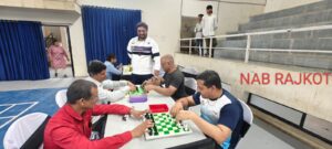 The photo is taken in an indoor sports hall. Multiple tables are set up where visually impaired individuals are intensely engaged in tactile chess games. They are using specially designed chess boards with raised black squares and distinguishable chess pieces for accessibility.