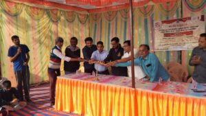 The image shows a group of men on a stage participating in a ceremonial event. They are collectively lighting a traditional brass lamp, which is a common practice in Indian culture to signify the beginning of an auspicious event or program.